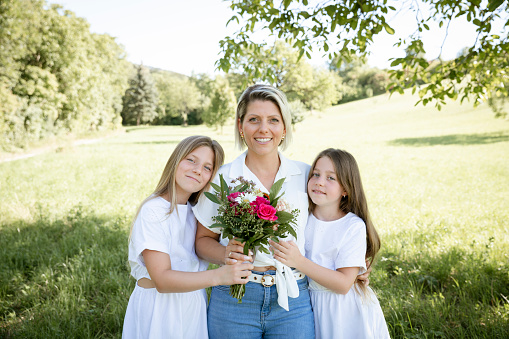 pretty young blonde mother getting bouquet of flowers from her two daughters for mother's day, birthday, valentines day outdoor in agreen meadow