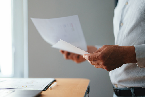 Close up photo of man hands holding business report while standing at office desk with a laptop computer.