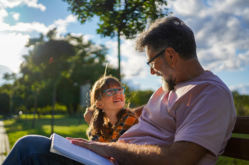 Grandfather and granddaughter outdoors reading books together at a park or back yard setting. Latin and Caucasian descent family.