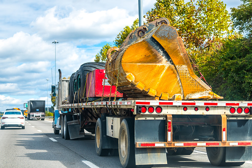 Closeup of industrial cargo freight truck trailer shipping hauling excavator machine equipment part of scoop bucket on highway road in traffic