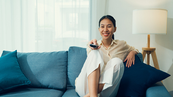 Beautiful Asian young woman on sofa click remote controller change channel on television choose favorite comedy or sitcoms in streaming online VOD, video on demand at home. Leisure activity concept.