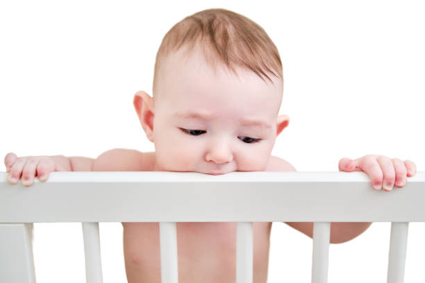 Baby gnaws at the edge crib during teething itching, isolated on a white background. Funny child scratching his teeth on the rail bed, age six months