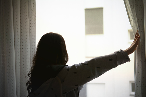 Rear view of young woman waking up in the morning, starting the day right. Looking through window