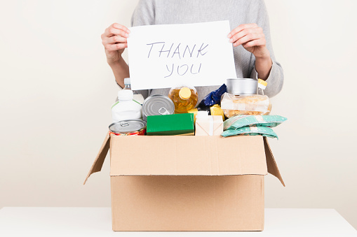 Female hands holding cardboard box with grocery products and card with inscription Thank you. Volunteer collecting food into box. Donations, charity, food bank, help for poor families.