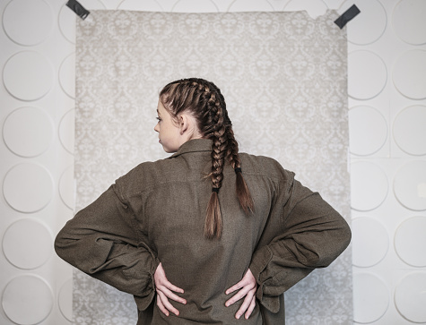 Studio Portrait of a girl actor in costume from the back. She is dressed in raggedy clothes of a poor person and has braided hair. Close up image in front of the damask pattern backdrop.