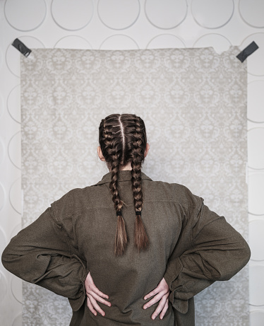 Studio Portrait of a girl actor in costume from the back. She is dressed in raggedy clothes of a poor person and has braided hair. Close up image in front of the damask pattern backdrop.