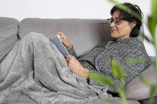 mature woman of middle eastern ethnicity happy and smiling brunette. she is reading a book on the sofa at home and covered with a warm blanket. concept of wellness and free time for oneself.