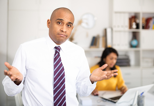 Puzzled and confused office employee making helpless gesture, standing on background with busy female colleague