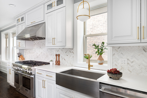 A luxurious white and grey kitchen with a large island, stainless steel Thermador appliances, and gold hardware, lights, and faucet.