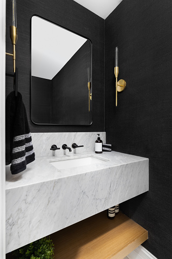 A bathroom with black wallpaper, gold sconces, and a floating marble sink and countertop.