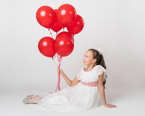 Beautiful girl dressed in long white dress holding lot of party red balloons in hand, looking up at balls. Studio shot of lying down girl of 10 years old on white background. Part of photo series.