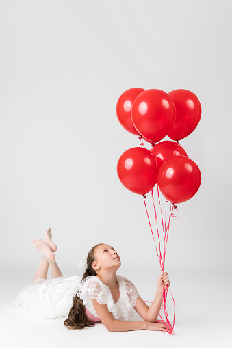 Beautiful happy girl on birthday holiday party, lying down and holding lot of red balloons in hand, looking up at balls. Studio shot of little Caucasian girl on white background. Part of photo series.