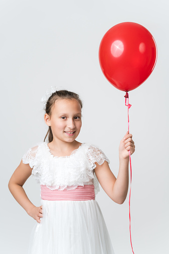 Portrait of girl in white dress holding red air balloons in hand, happy smiling looking at camera. Front view studio shot of Caucasian model ten years old on white background. Part of photo series