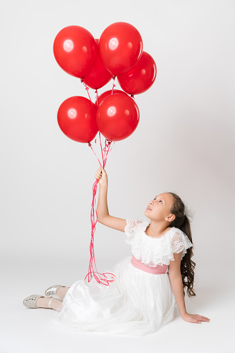 Smiling girl dressed in long white dress holding large bunch of party red balloons in hand, looking up at balls. Studio shot of sitting Caucasian girl of 10 years old on white background. Part of photo series