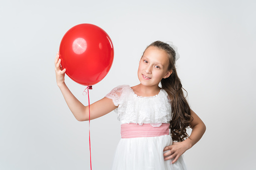 Portrait of happy girl in white dress holding one red balloon in hand, smiling and looking at camera. Studio shot of attractive Caucasian model 10 years old on white background. Part of photo series