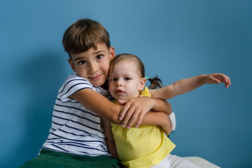 Two kids preschool boy hugs his little sister siblings in front of blue wall indoor portrait childhood growing up concept copy space front view