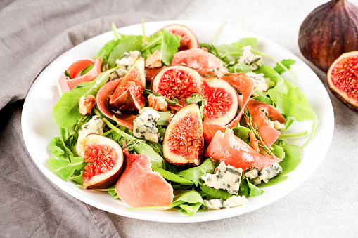 Healthy salad with fresh fig, blue cheese, prosciutto, arugula and spinach on white table background with ripe figs around. Selective focus