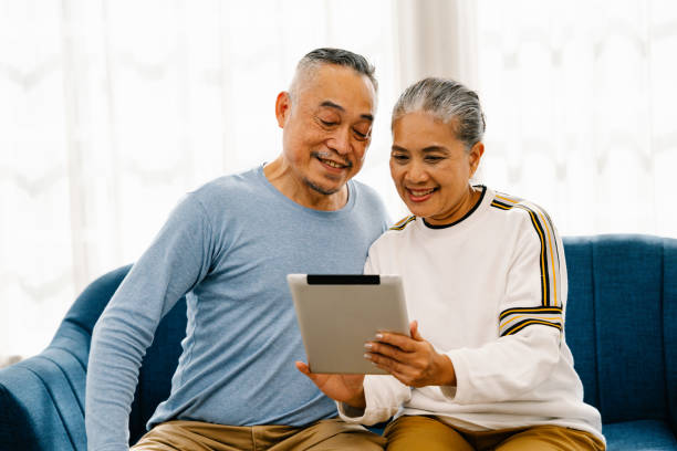 istock Smiling senior couple sitting on the couch looking at tablet computer, Asian retirees use contemporary technology 1449496383