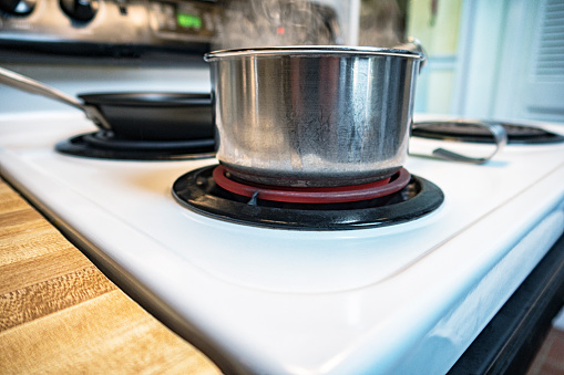 Close-up of a red hot electric burner heating under a well-used aluminum and stainless steel metal saucepan pot of boiling water steaming on an also well-used vintage electric range stove top.