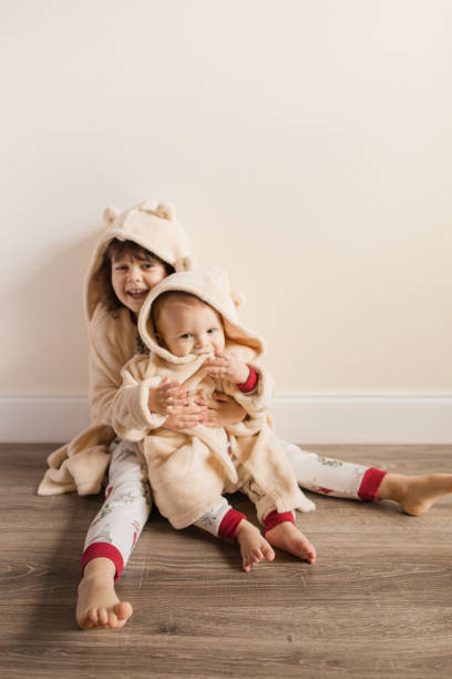 Three-Year-Old Toddler Girl Sitting on the Floor Holding Her 11-Month-Old Baby Brother as They Wear Matching Simple Christmas Pajamas & Fluffy Cream-Colored Bear-Themed Robes For a Cozy Christmas Season At Home in December 2022 Three-Year-Old Toddler Girl Sitting on the Floor Holding Her 11-Month-Old Baby Brother as They Wear Matching Simple Christmas Pajamas For a Cozy Christmas Season At Home in December 2022 3 6 months stock pictures, royalty-free photos & images