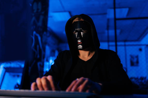 Dangerous unrecognizable hacker man wearing in black mask and sweatshirt hacking online web site or engaging hacking into security systems