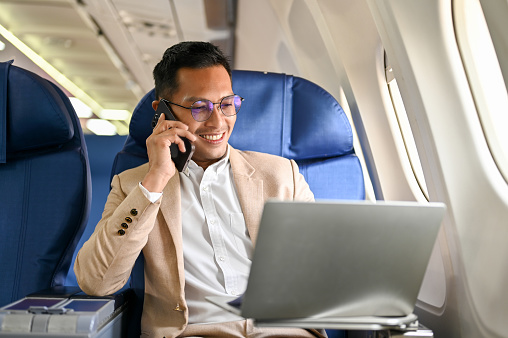 Successful and smart Asian businessman looks at his laptop screen, on the phone with his business client during the flight for an overseas business meeting.