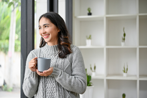 Beautiful and happy Asian woman in cozy sweater holding a cup of coffee, smiling, looking out the window, daydreaming about success and happiness.