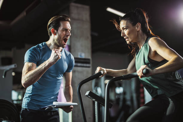 trainer feuert athletische frau auf dem heimtrainer in einem fitnessstudio an. - spinning instructor exercising gym stock-fotos und bilder