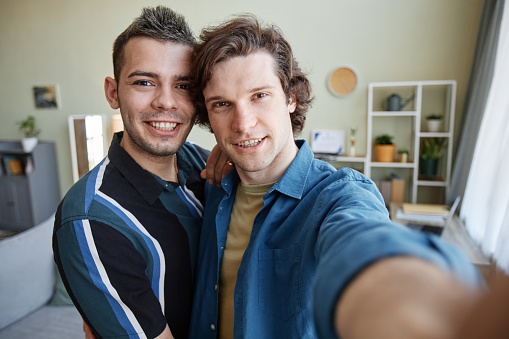 Portrait of young gay couple taking selfie photo in new home together, camera POV