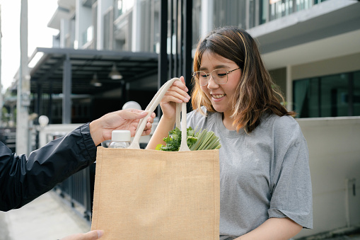 Asian woman receiving vegetables and prawns from delivery man after ordering from an online grocery store to cook Thai food at home.