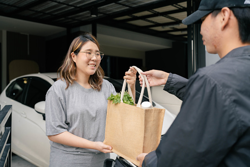 Asian woman receiving vegetables and prawns from delivery man after ordering from an online grocery store to cook Thai food at home.