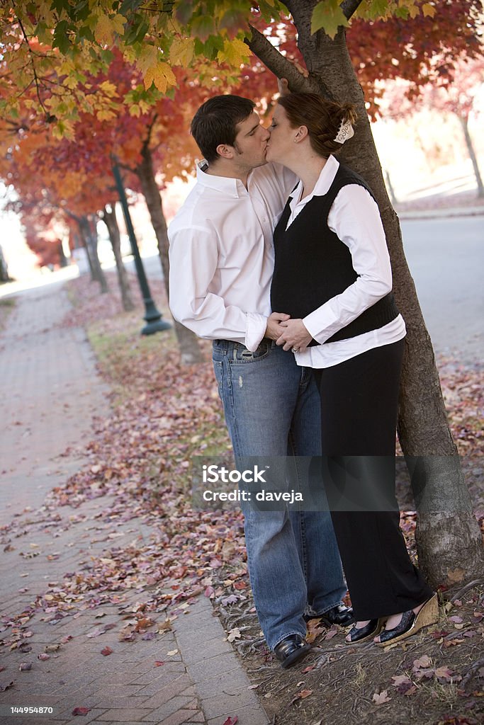 Kissing on Fall Day A young couple kisses under a row of beautiful fall trees. They hold hands on her pregnant belly. Abdomen Stock Photo