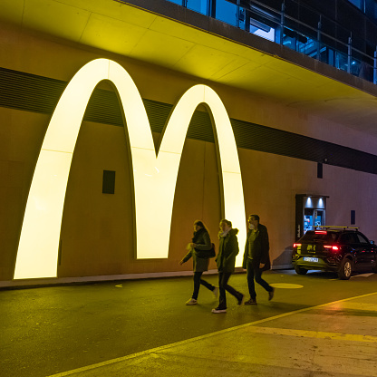 Ondara, Alicante province, España -  December 5th, 2022: McDonald's big logo illuminated at night in the drive through service with a car waiting and people passing through. McDonald's is the world's largest chain of hamburger fast food restaurants.