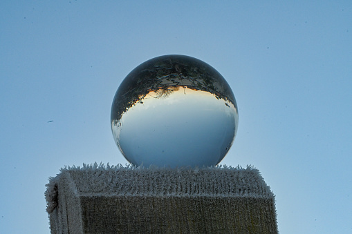 A glass crystal ball sitting on top of a frost covered fence post, with a clear blue sky background, reflecting upside down the surrounding landscape environment