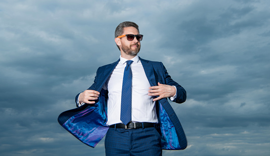 Businessman wearing open suit jacket fluttering in wind. Grizzled businessman in sunglasses. Businessman sky background.