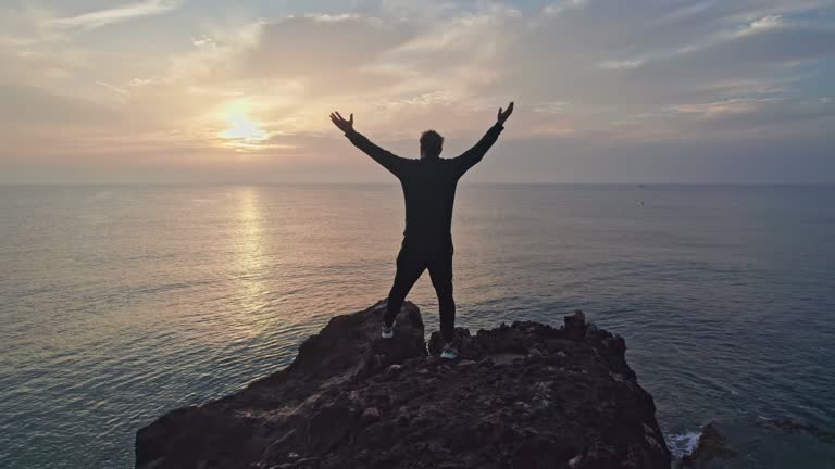 Successful man raising his hands in success gesture. Happiness of reaching the summit. Silhouette on the rock by the sea.