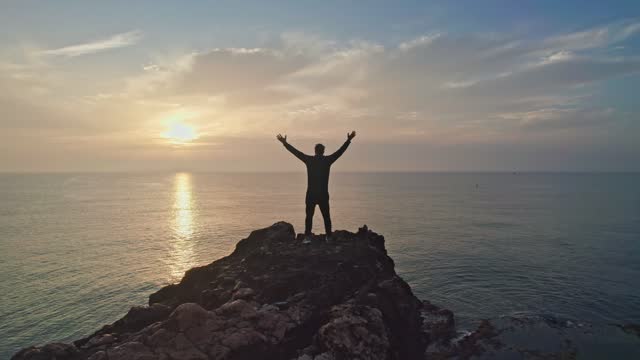 Successful man raising his hands in success gesture. Happiness of reaching the summit. Silhouette on the rock by the sea.