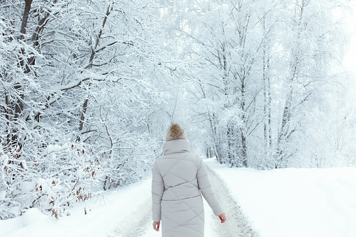 Woman from behind in warm knitted hat walking through a snowy road, white wooded path in a forest in winter time