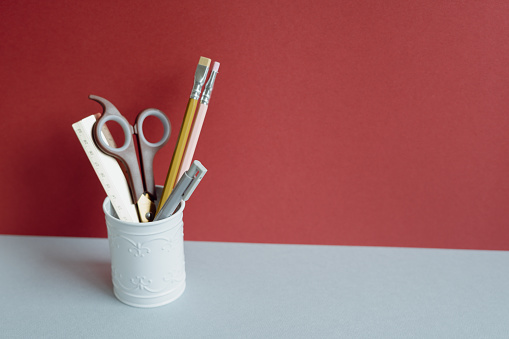 Ruler, scissors, colored pencil, pen in pencil holder case on gray desk. red wall background. copy space