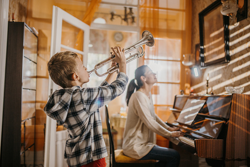 Young boy playing a trumpet, while his mother is playing piano, they are playing music together at home.