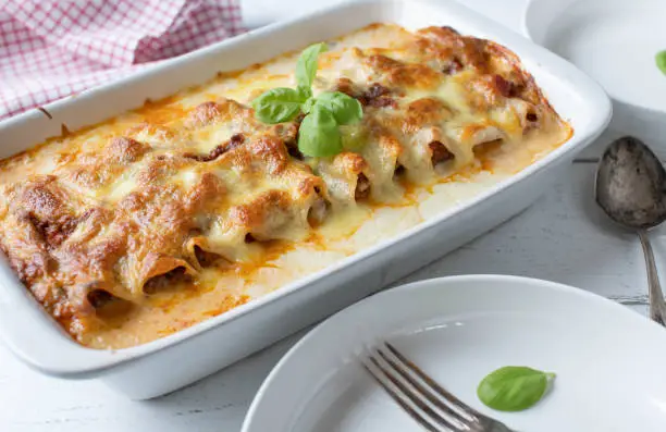 Italian cusine. Traditional noodle casserole or lasagna with cannelloni noodles. Filled with bolognese sauce and topped with bechamel sauce and mozzarella cheese. Served in a white casserole dish on white wooden background. closeup