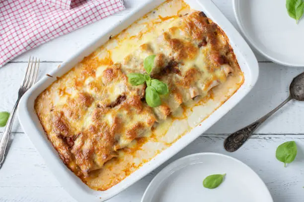 Traditional italian pasta casserole with stuffed cannelloni with bolognese sauce. Topped with bechamel sauce and mozzarella cheese topping. Served in a white casserole dish on white wooden background. Table top view