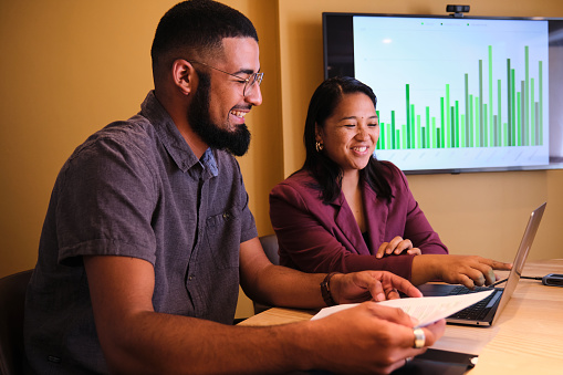 Two diverse young businesspeople laughing while going over paperwork and working on a laptop in an office boardroom