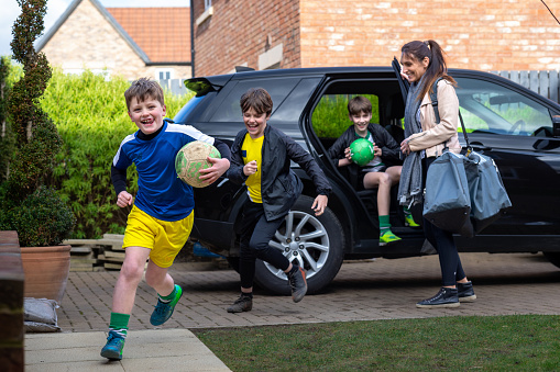 A three-quarter length of a mother opening the car door for her children on the drive. The children are running out of the car wearing football attire.