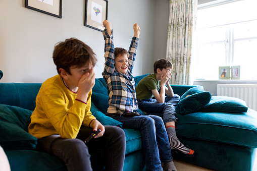 A three-quarter length shot of three brothers playing video games together all sitting on a blue sofa. The young boy in the middle has his hands raised smiling.