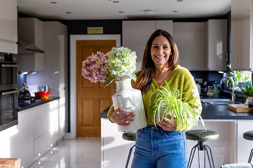 A three-quarter length shot of a mature female standing in her kitchen holding flowers and a houseplant. She is smiling into the camera wearing casual clothing.