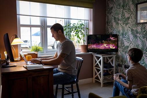 A three-quarter length shot of a father working from home on his laptop to the left and his son playing a video game to the right. They are both concentrating on what they are doing.