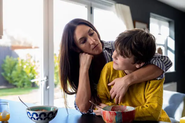 A waist-up shot of a mother reaching her arm around her son next to the kitchen counter looking unhappy with her son. They are wearing casual clothing.