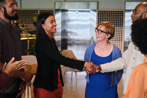 istock Laughing businesspeople shaking hands while standing with coworkers in an office 1449604950