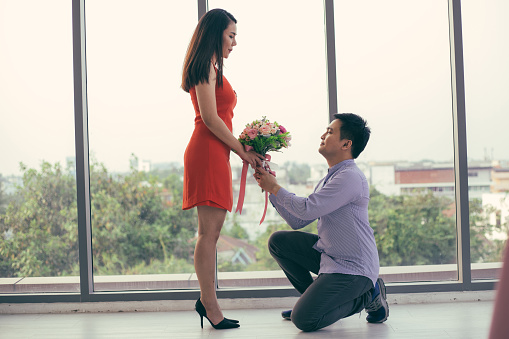 Asian man standing on one knee giving flower to propose his girlfriend on Valentine's day. couple having romantic dating for anniversary or birthday, Love and marriage concept